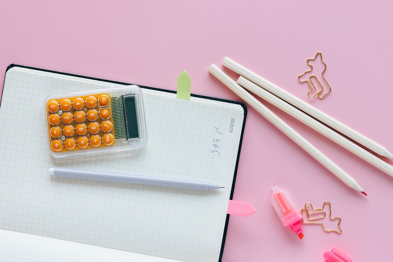 Home Top view of office supplies including a calculator, pencils, and clips on a pink background.