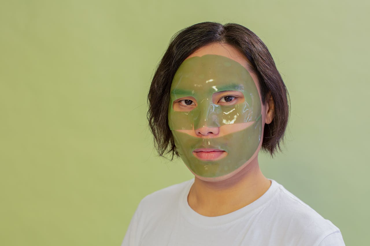 About Adult wearing a green facial mask on a light green background, promoting skincare and wellness.